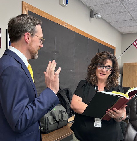 A man in a blue suit raises his right hand and takes an oath of office. A woman on the right with dark hair holds a book while reading the oath to him.