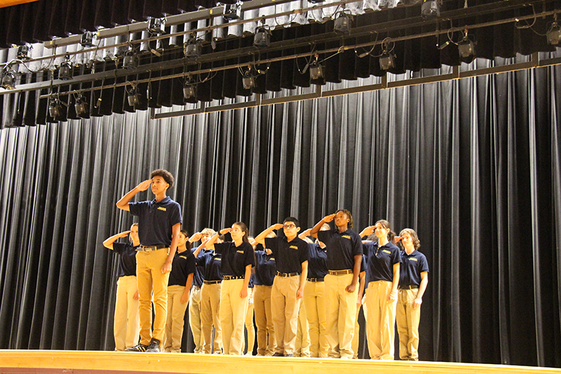 A group of about 20 high school students, all dressed in khaki pants and navy blue shirts, stand in three lines with one student in the front, all saluting the audience from a stage.