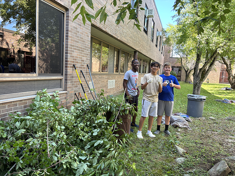 Three middle school age boys stand outside of a building giving thumbs up. They are doing yardwork, with rakes and shovels nearby and bins filled with greenery. 