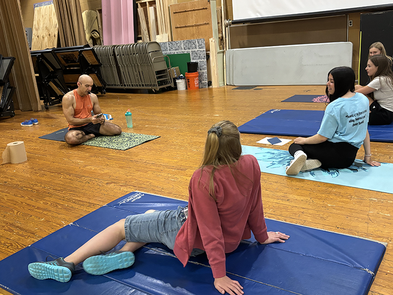 A man sits on a yoga mat and in front of him are middle school kids also on mats.