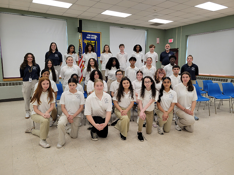 A group of 30 high school kids in white shirts and khaki pants, stand and kneel for a group picture. There are four adults in navy blue shirts with them.