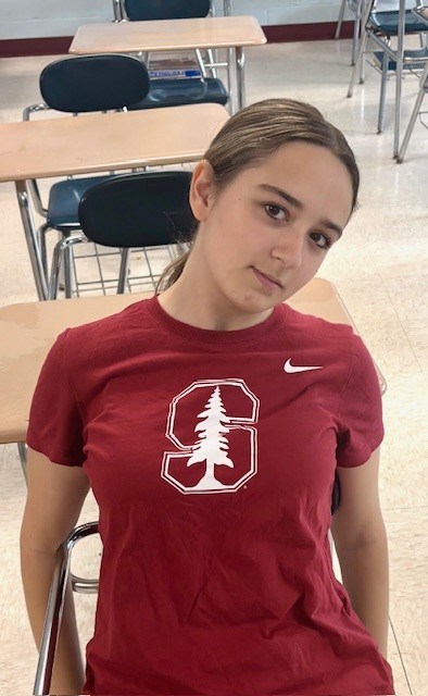 A young woman with long dark hair pulled back into a ponytail, sits at a desk in a classroom. She is wearing a red tshirt with the Stanford University logo on it - an S with a pine tree in the center.