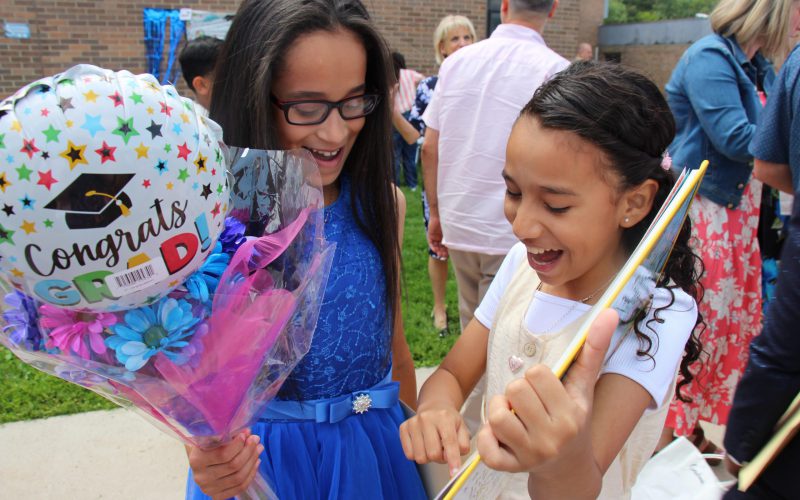 Two girls look excitedly at a yearbook. One is holding a congrats Grad balloon.