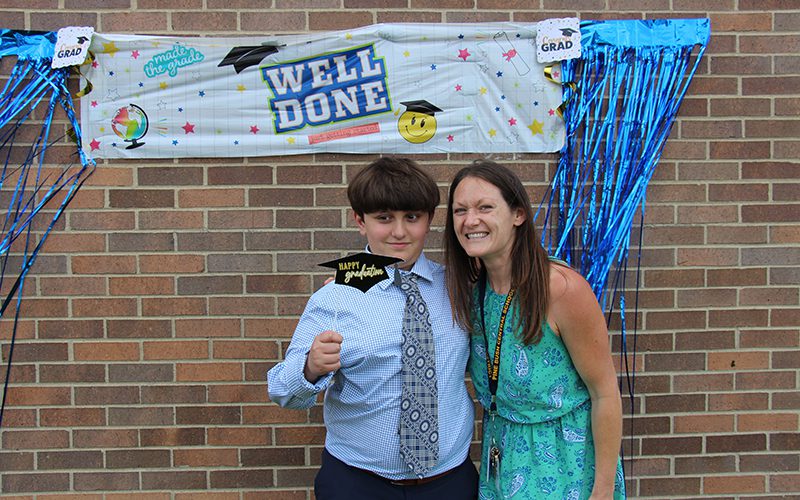 A sign in th ebackground says well done. A fifth-grade boy in a blue shirt and tie holds a little graduation cap on a stick that says Happy graduation. With him is a woman with long dark hair smiling.
