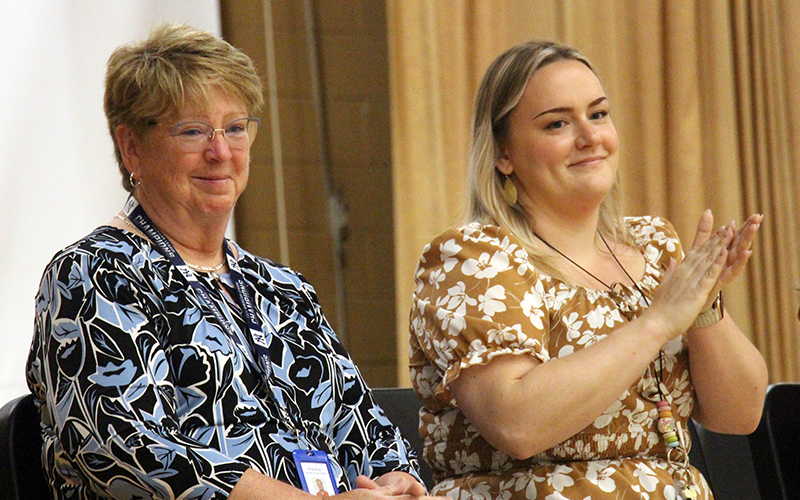 Two women sit on a stage and smile clapping.