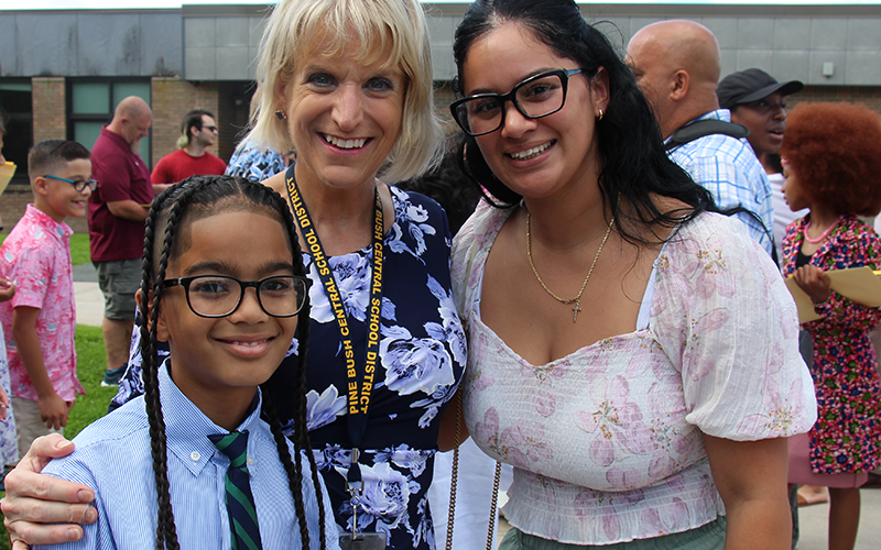 Two women stand smiling with a fifth-grade boy who is wearing a blue shirt and tie, glasses and has long dark braids.