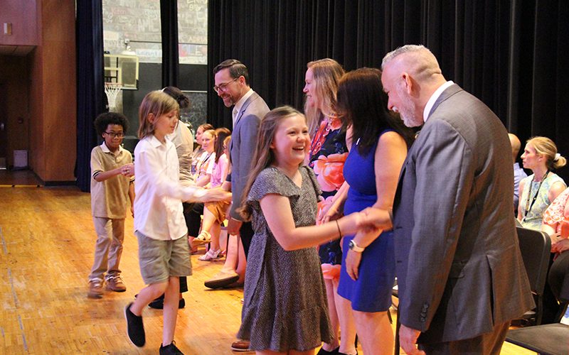 A girl smiles broadly as she shakes hands with a man on a stage. Two boys behind her are also shaking hands with adults.