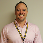 A man smiles. He is wearing a checked button down shirt, has short dark hair and light facial hair.