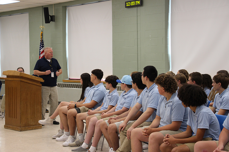 A group of high school kids sit and listen to a an speaking.