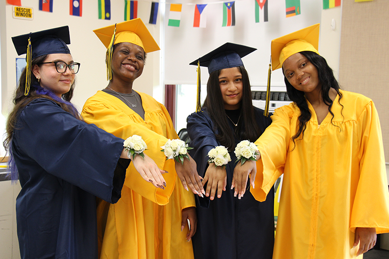Four high school senior girls, all wearing either a blue or gold graduation cap and gown, hold their arms out to show flowers they are wearing on their wrists. They are all smiling.
