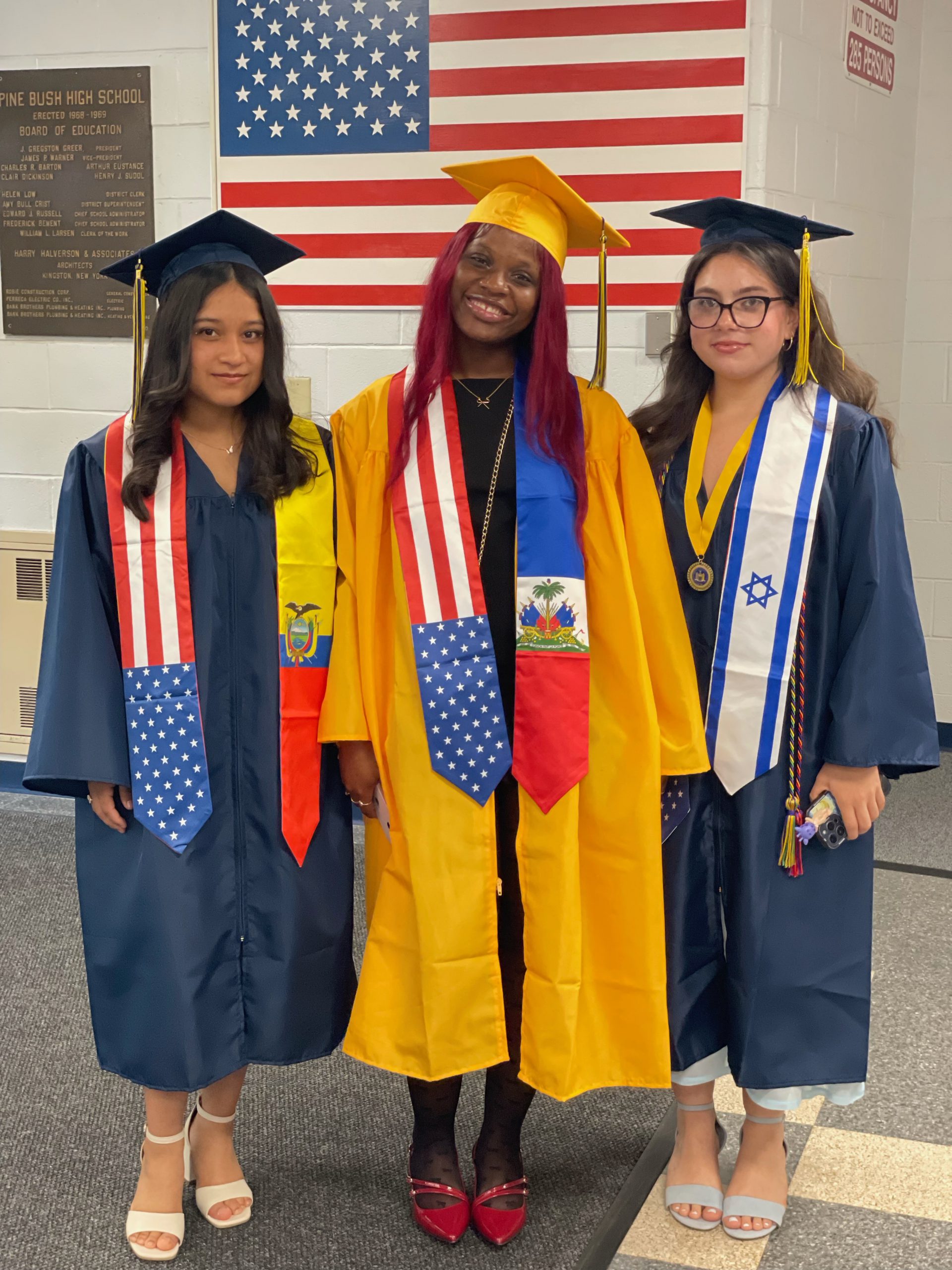Three girls stand in their blue and gold caps and gowns with stoles around their shoulders. One side is the American flag and the other is the flag of Haiti, Ecuador or Israel.