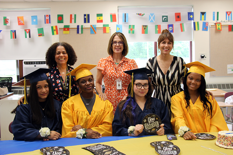 Four high school senior girls, dressed in blue or gold caps and gowns, sit at a table with graduation napkins in front of them. Behind them are three women standing and smiling.