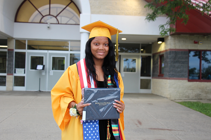 A young woman in a gold cap and gown holds her diploma in front of her. She has long dark hair and is smiling.