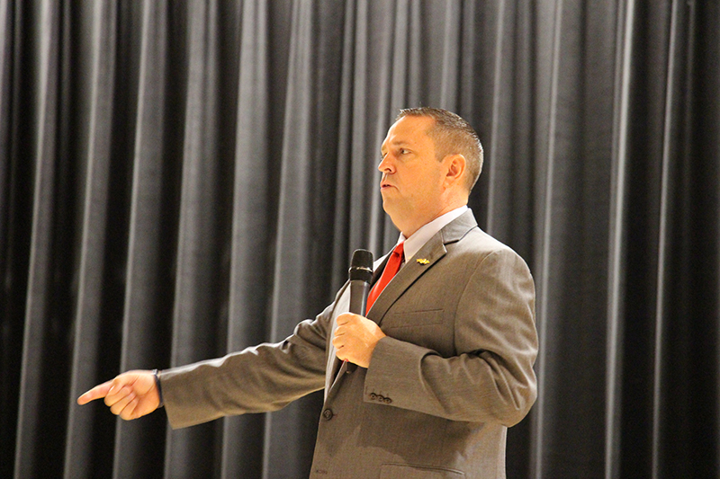 A man with short dark hair holds a microphone and speaks to a group of people from a stage. He is wearing a suit and tie.