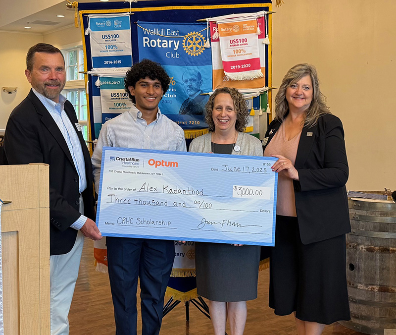 A young man with dark hair smiles and holds an oversized check. He is standing with two women on the right and a man on the left.