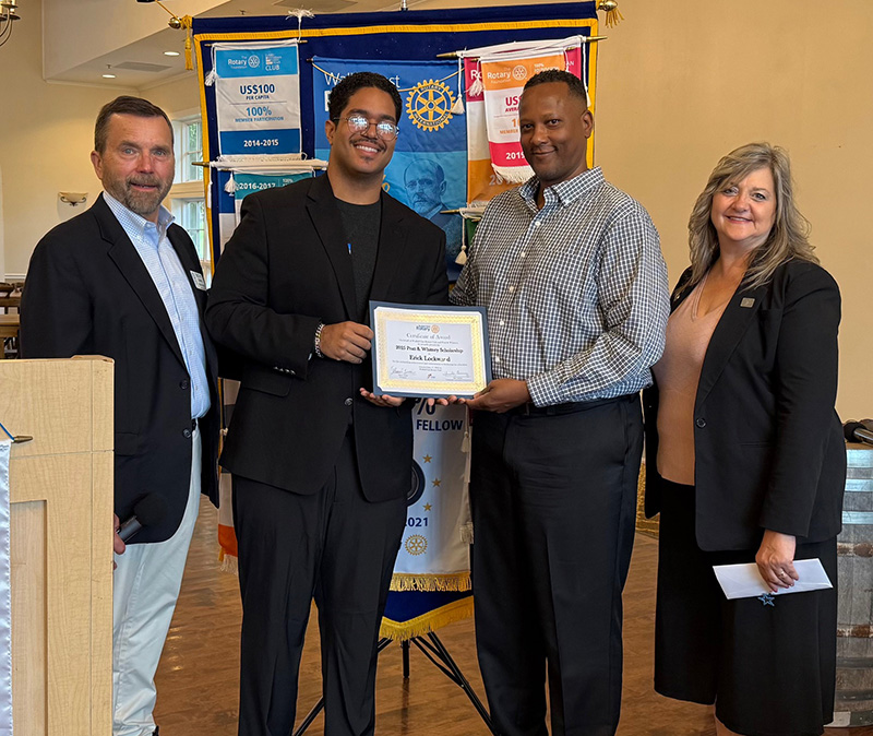 A tall young man with glasses and short dark hair smiles holding a plaque. He is surrounded by two men and one woman.