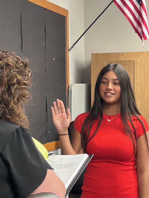 A young woman wearing a red dress holds her right hand up as she takes an oath of office. She is smiling.