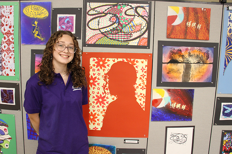 A high school girl stands in front of several pieces of art she made. She is wearing a purple shirt, glasses and has longer brown hair. She is smiling.
