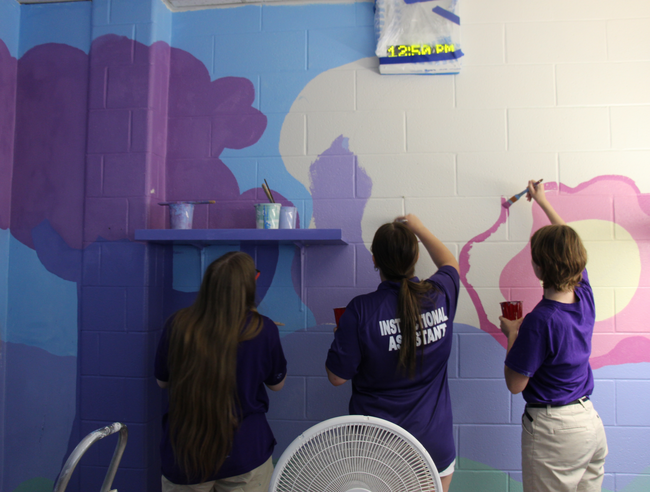Three high school art students paint a mural on a wall. They are using blue, different shades of purple and white.