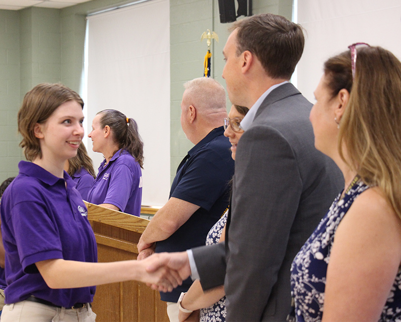 A young woman in a purple shirt smiles and shakes hands with a man. Others are waiting to shake her hand.