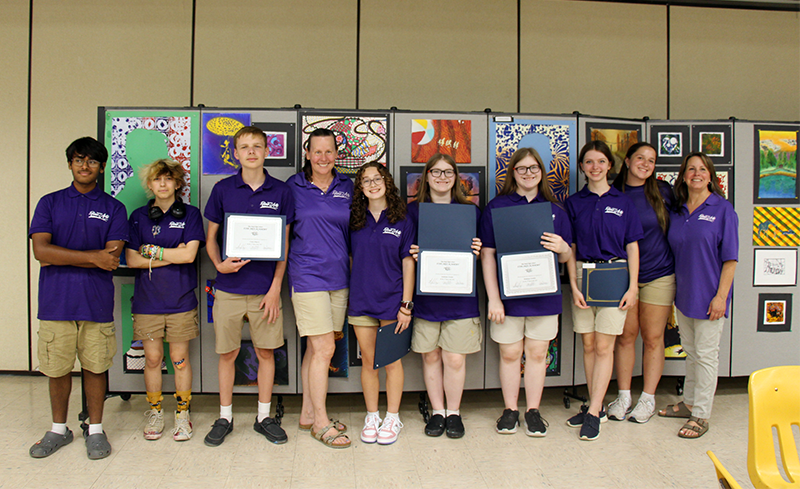 Two women and seven students stand smiling in front of a wall of art they created. Several are holding the certificates they just earned.