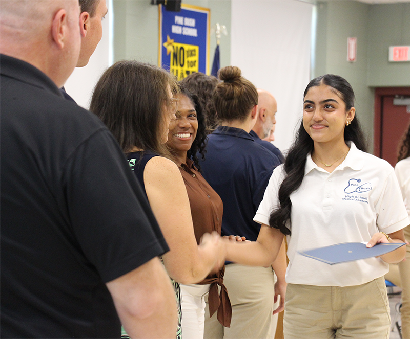 A young woman in a white shirt and long dark hair shakes hands with a woman.