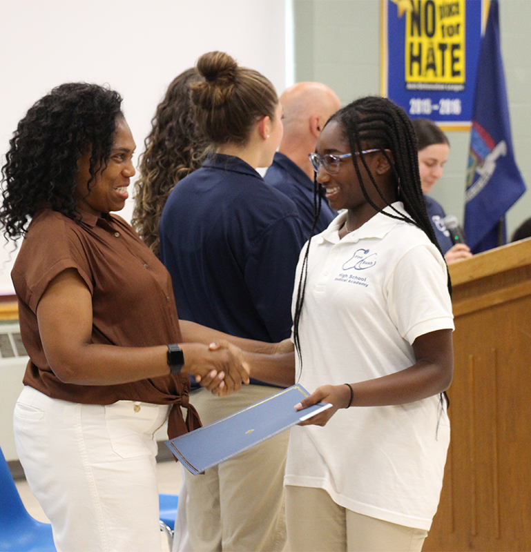 A high school girl in a white polo shirt smiles broadly as she shakes hands with a woman who is also smiling.