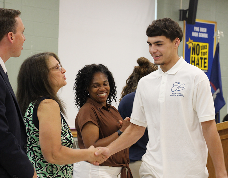 A high school age young man smiles as he shakes hands with a woman. Another woman smiles and looks on.