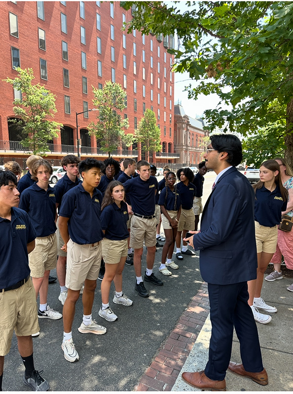 A large group of high school students wearing khaki shorts and navy blue shirts stand and listen to a man speaking to them. He is wearing a blue suit.