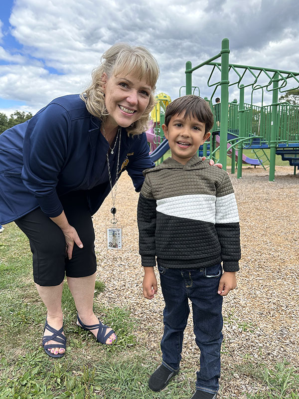 A woman with shoulder-length blonde hair smiles  standing with an elementary age boy on a playground.