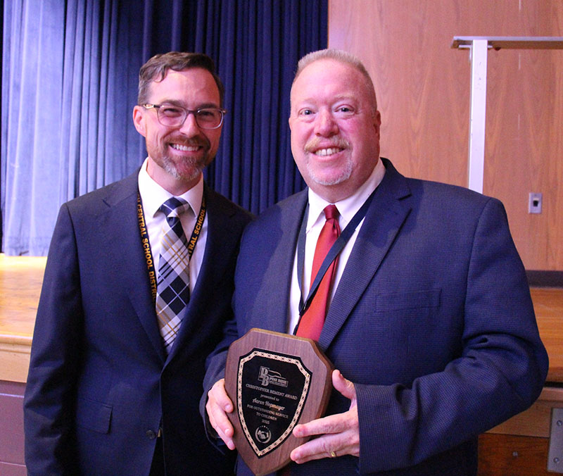 Two men stand together smiling, both in dark suits. The man on the right is holding an award.