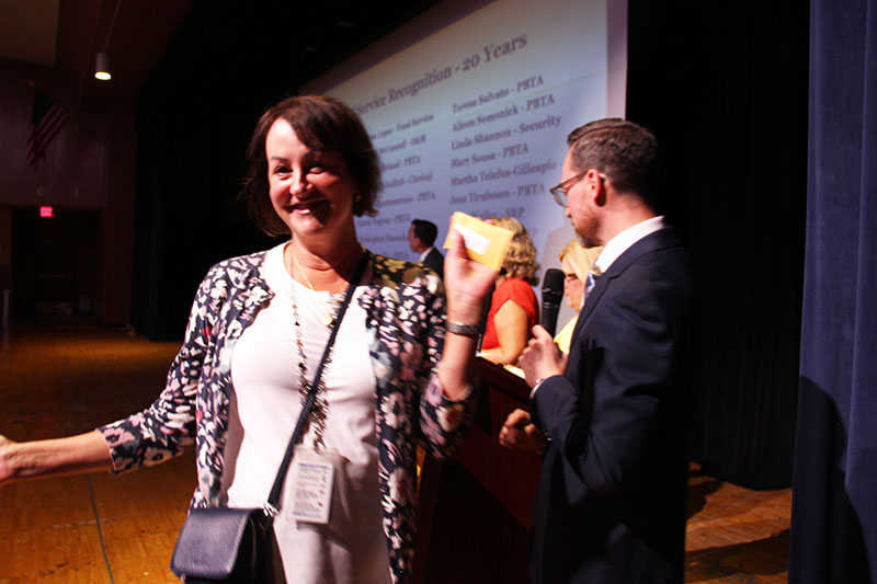 A woman with short dark hair smiles broadly and holds up an envelop.