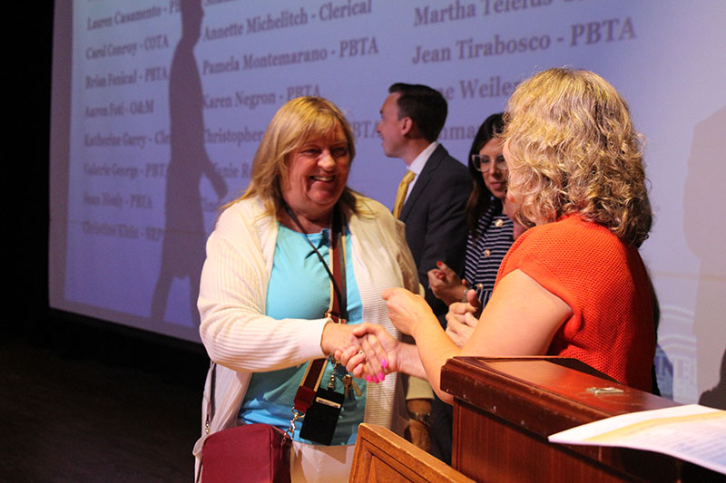 A woman in a blue shirt and tan sweater shakes hands with a woman with blonde hair.
