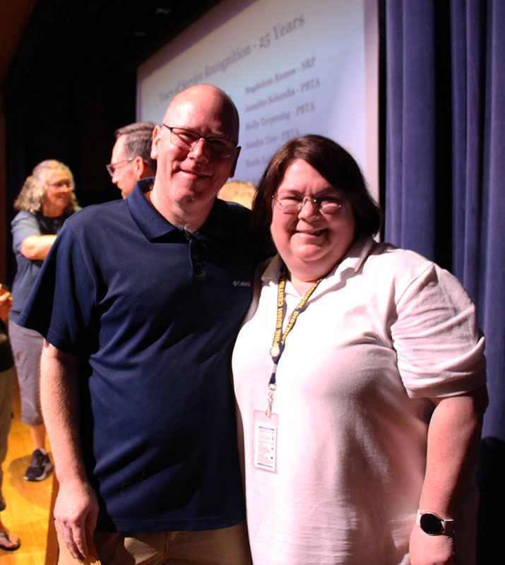 A man and woman stand together smiling, with arms around each other. 