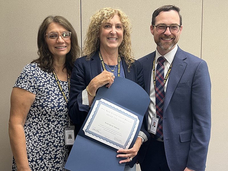 Two women and a man smile. The woman in the center ha slonger blonde hair and is holding a certificate.