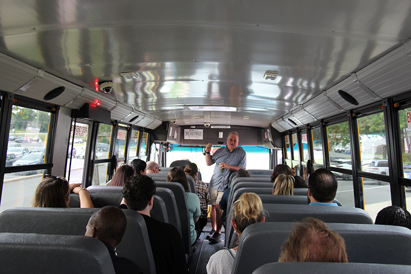 Inside a school bus, adults are sitting in the seats. At the front of the bus is a man speaking to everyone.
