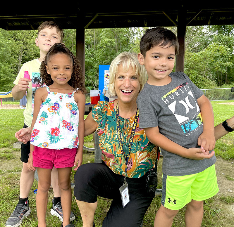 A woman kneels down with three kids smiling. Some of the kids have popsicles.
