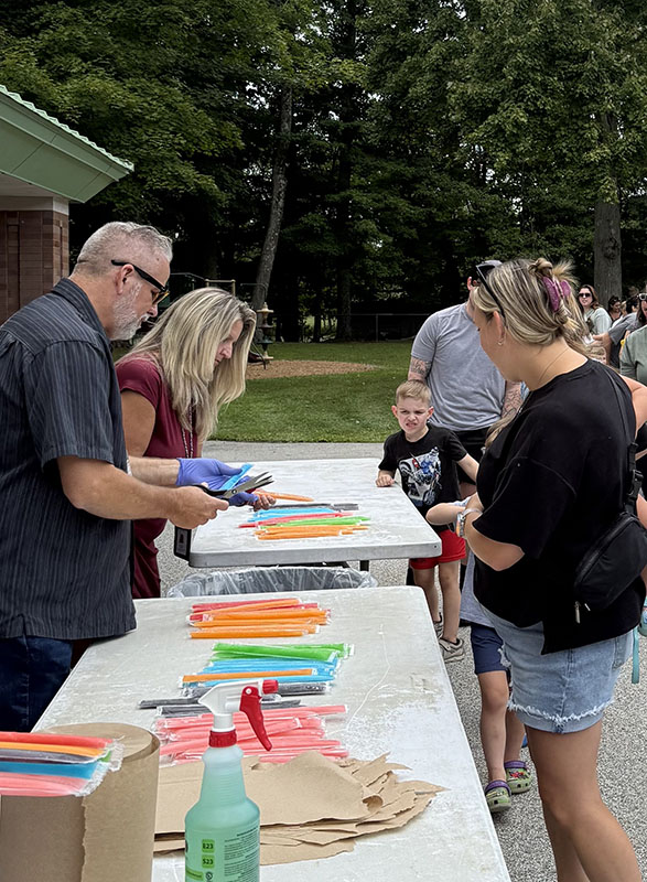 A man and woman stand behind a table handing out popsicles to adults and children.