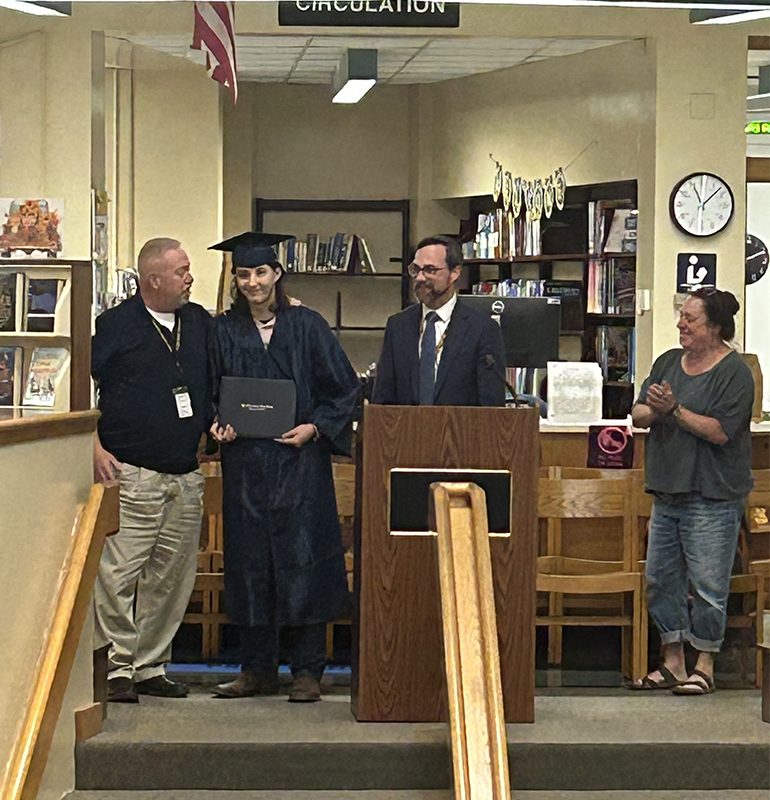 a man stands at a podium speaking. There is another man on the left and a young man in a blue cap and gown. On his right is a woman clapping.
