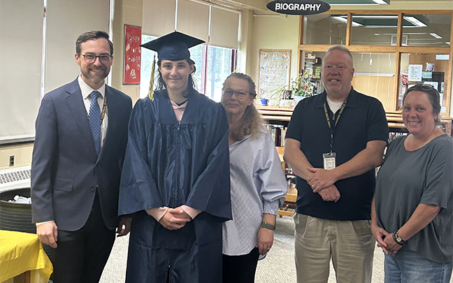 A group of five people stand together smiling. From left, a man in a suit and tie smiling, a young man wearing a blue cap and gown, a woman in a white shirt, a man in blue shirt and khaki pants, and a woman in a blue shirt and jeans. They are all smiling.