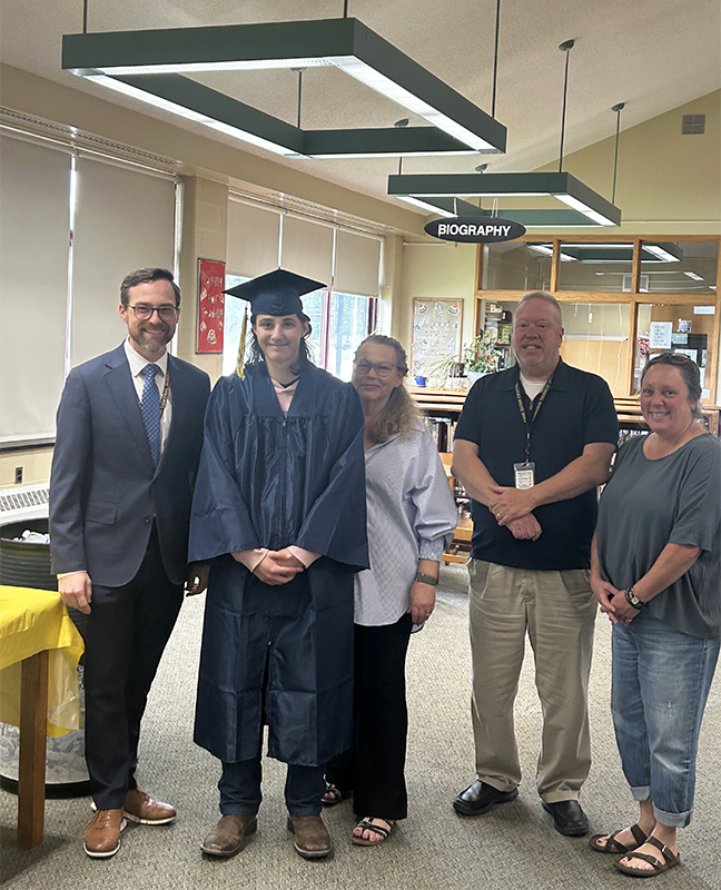 A group of five people stand together smiling. From left, a man in a suit and tie smiling, a young man wearing a blue cap and gown, a woman in a white shirt, a man in blue shirt and khaki pants, and a woman in a blue shirt and jeans. They are all smiling.