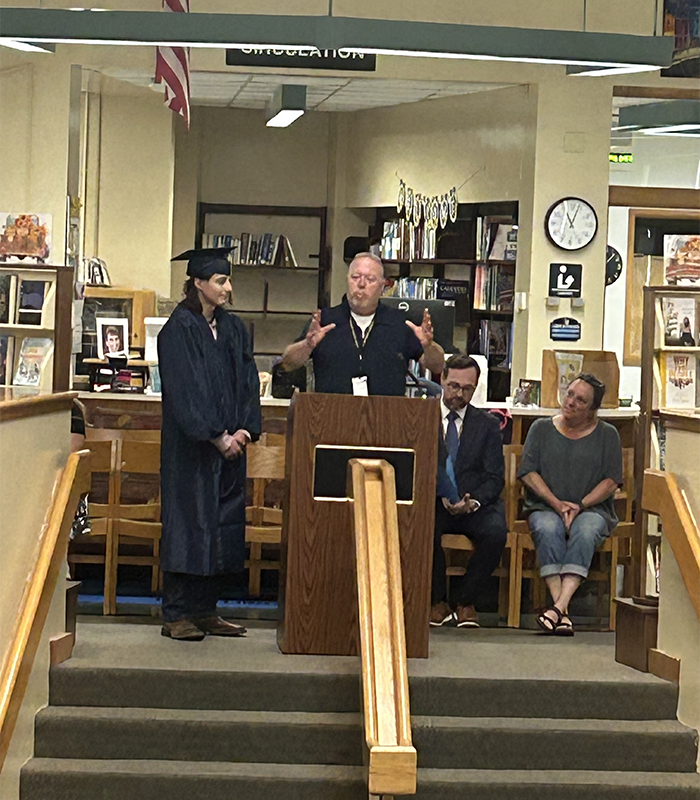 A man stands at a podium talking. Next to him is a young man with long dark hair, wearing a blue cap and gown.