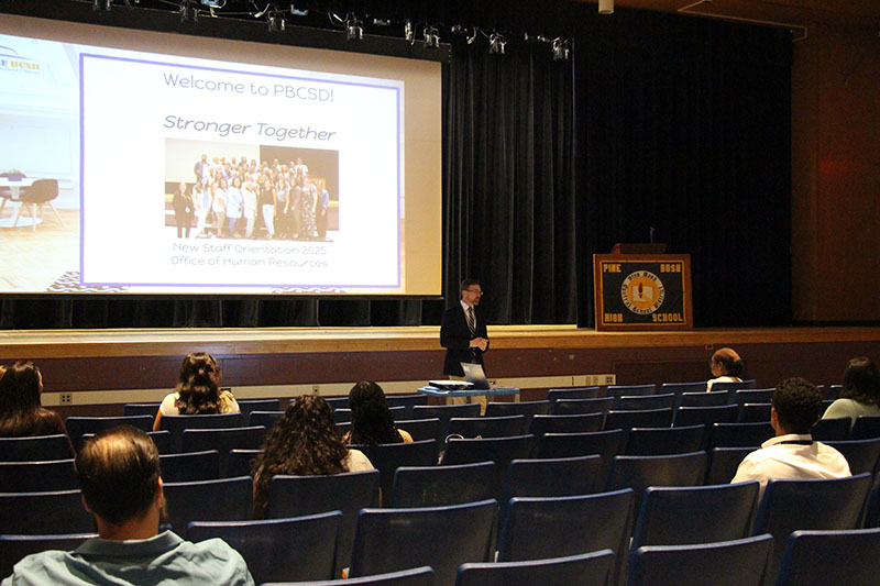 In an auditorium, a man stands in front of a group of people spread out in seats. Behind him is a screen that says Stronger Together.