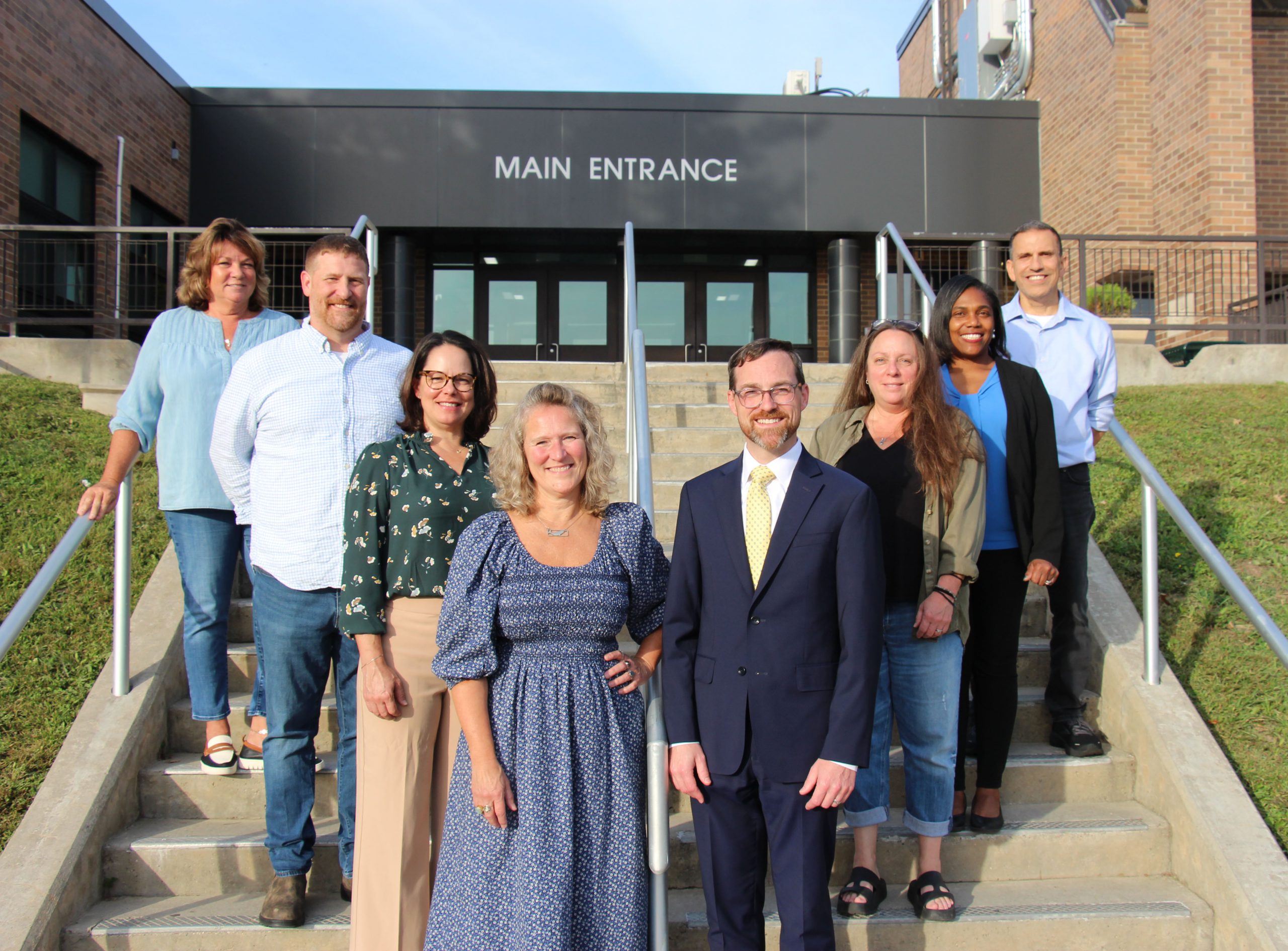 Eight adults - three men and five women - stand on a set of concrete steps in a v form. All are smiling. There is a brick school building behind them.