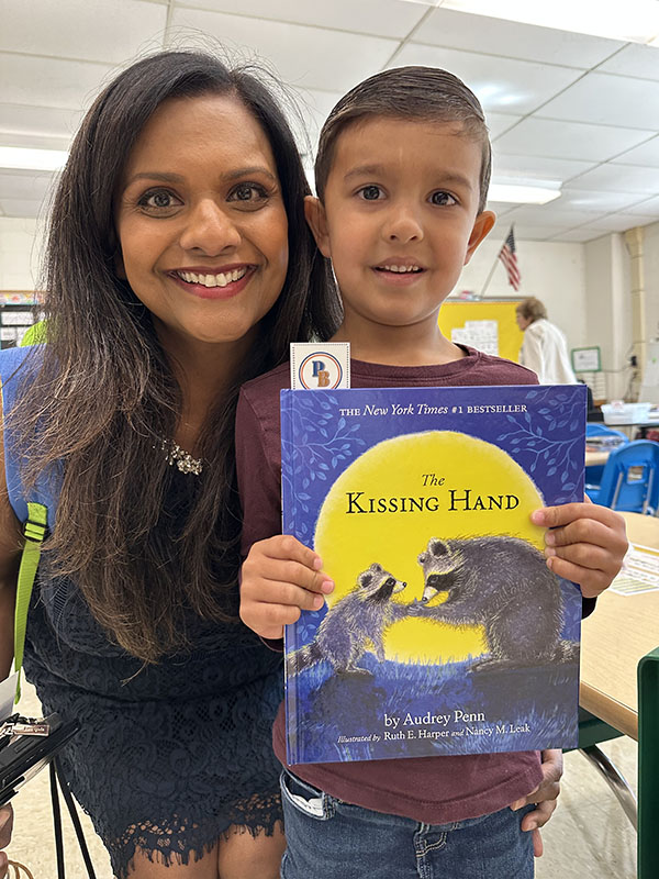A woman smiles. She has long dark hair. Next to her is a little boy also smiling. He is holding a book called The Kissing hand.