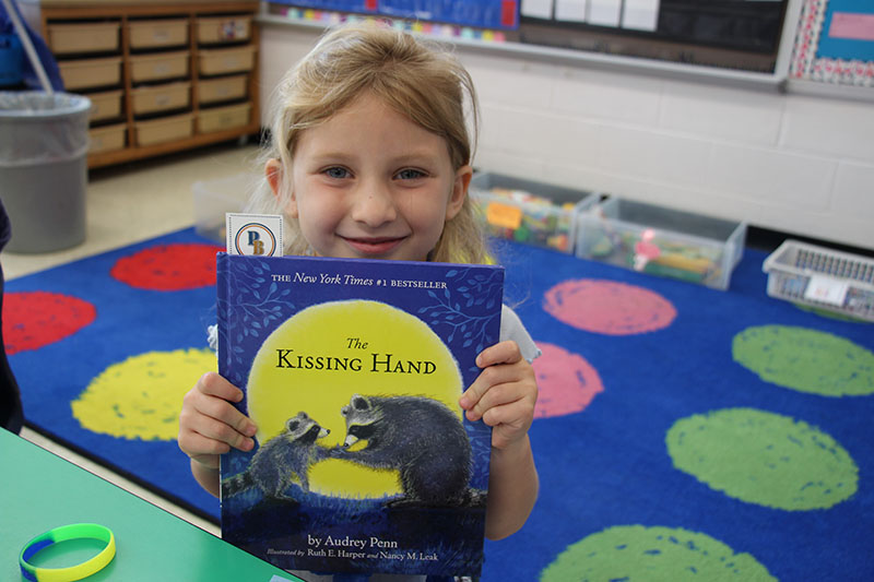 A little girl with blond hair smiles and holds a book called The Kissing Hand.