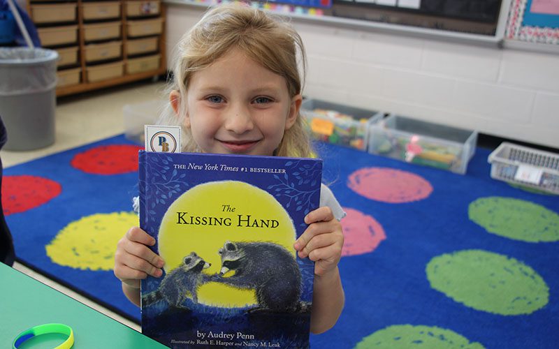 A little girl smiles and holds a book called The Kissing Hand.