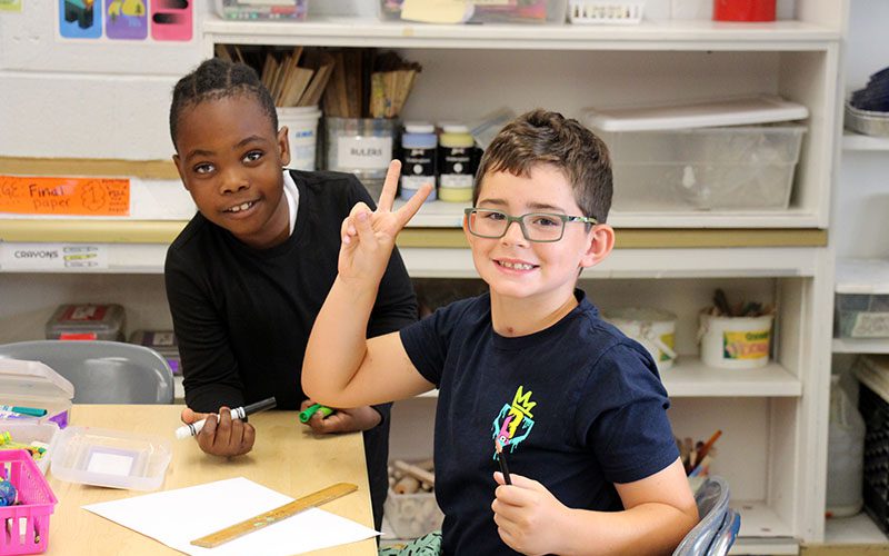 Two second-grade boys smile as they work on art projects. The boy on the right has short hair and glasses and is giving a peace sign. The boy on the left is holding a marker and has short dark hair.