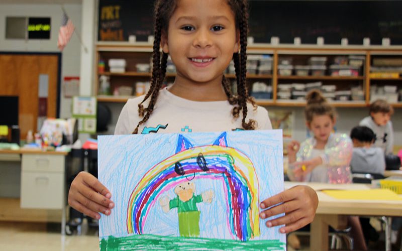 A young elementary aged girl with braids holds up her art project which is a very colorful picture of someone under a rainbow.
