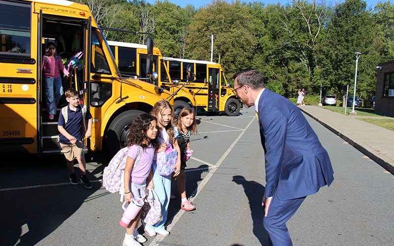 Elementary students get off a bus and stop to talk to a man in a suit who is crouched down to talk to them.